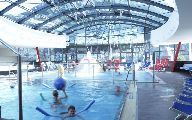 Innenpool mit im Wasser spielenden Kindern, umgeben von Liegestühlen und umschlossen von einem Glasdach und Glaswänden.