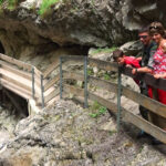Eine Familie steht auf einem Holzsteg mit Metallgeländer, der entlang einer felsigen Schlucht oberhalb eines Baches gebaut und von steilen Felswänden umgeben ist.