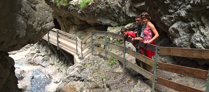 Eine Familie steht auf einem Holzsteg mit Metallgeländer, der entlang einer felsigen Schlucht oberhalb eines Baches gebaut und von steilen Felswänden umgeben ist.