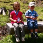 Zwei kleine Kinder mit Medaillen sitzen auf einer Holzbank im Freien, mit Gras und Baumstümpfen im Hintergrund.