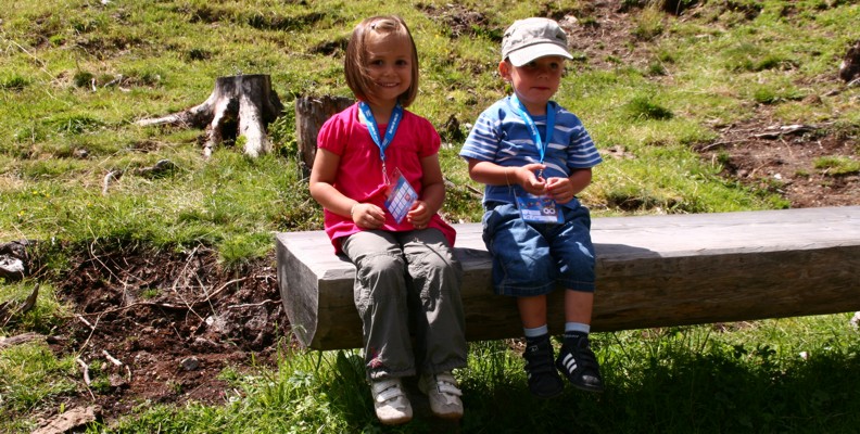 Zwei kleine Kinder mit Medaillen sitzen auf einer Holzbank im Freien, mit Gras und Baumstümpfen im Hintergrund.
