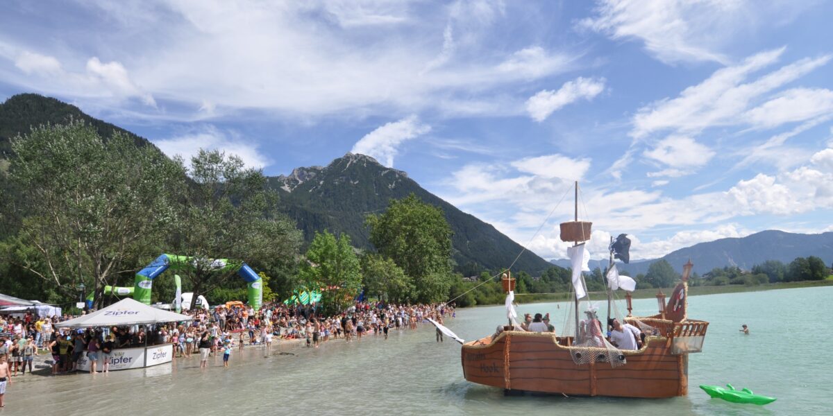 Eine große Menschenmenge versammelt sich am Ufer eines Sees mit Bergen im Hintergrund, während ein hölzernes Piratenschiff in der Nähe des Ufers im Wasser schwimmt.