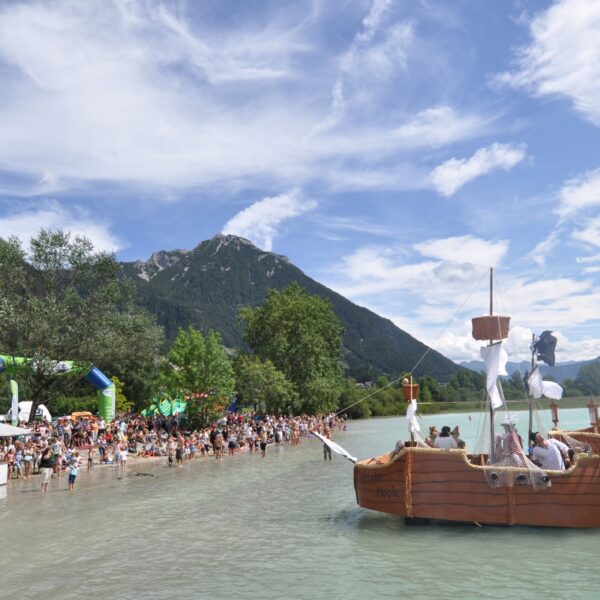 Eine große Menschenmenge versammelt sich am Ufer eines Sees mit Bergen im Hintergrund, während ein hölzernes Piratenschiff in der Nähe des Ufers im Wasser schwimmt.