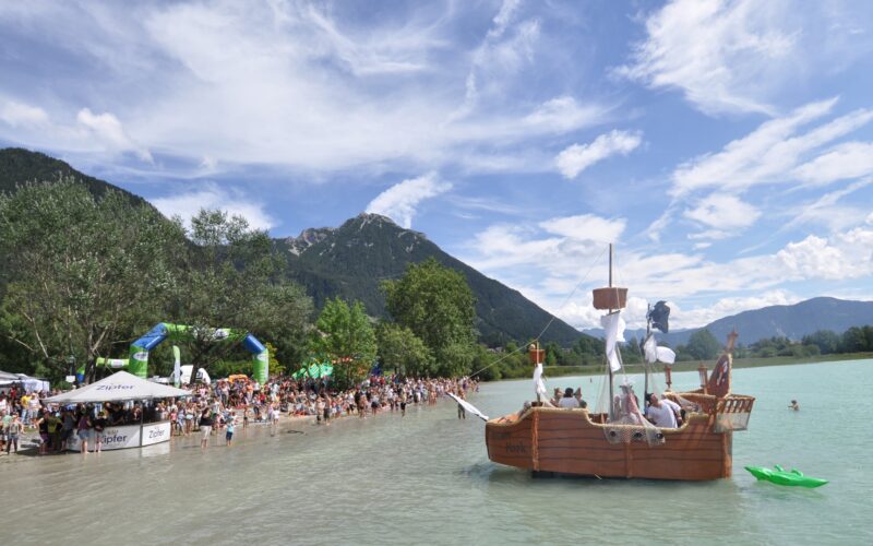 Eine große Menschenmenge versammelt sich am Ufer eines Sees mit Bergen im Hintergrund, während ein hölzernes Piratenschiff in der Nähe des Ufers im Wasser schwimmt.