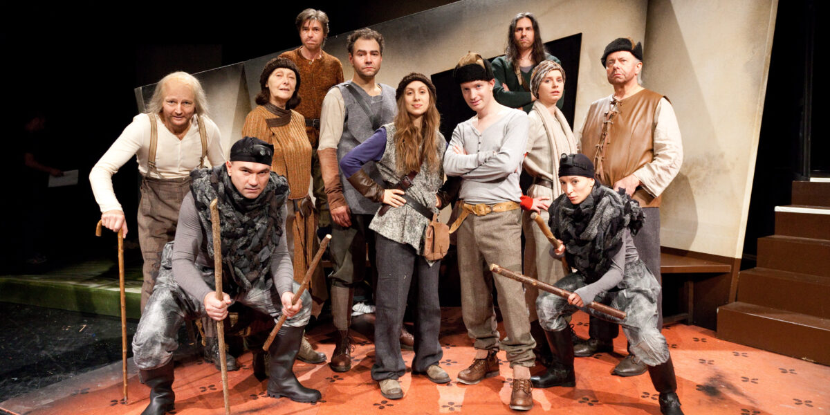 Eine Gruppe von Menschen in mittelalterlichen Kostümen posiert auf einer Theaterbühne mit Requisiten und bereitet sich wahrscheinlich auf oder nach einer Theateraufführung vor.