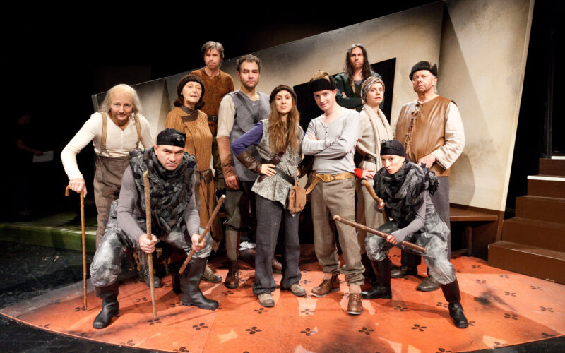 Eine Gruppe von Menschen in mittelalterlichen Kostümen posiert auf einer Theaterbühne mit Requisiten und bereitet sich wahrscheinlich auf oder nach einer Theateraufführung vor.