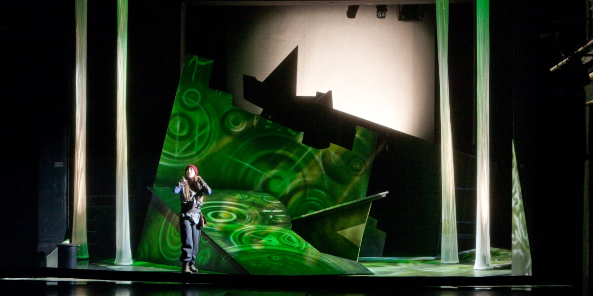 Ein Performer steht auf einer modernen, abstrakten Bühne mit grüner Beleuchtung, geometrischen Versatzstücken und vertikalen Lichtsäulen, die Kreise projizieren.