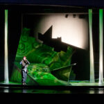 Ein Performer steht auf einer modernen, abstrakten Bühne mit grüner Beleuchtung, geometrischen Versatzstücken und vertikalen Lichtsäulen, die Kreise projizieren.