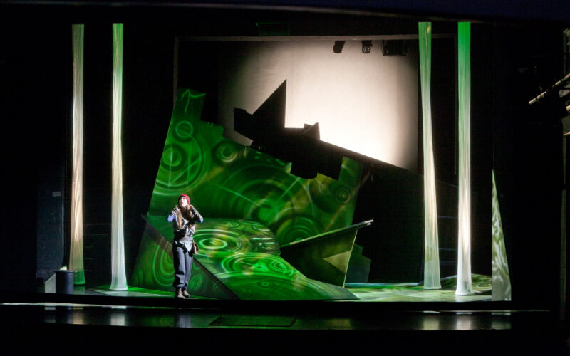 Ein Performer steht auf einer modernen, abstrakten Bühne mit grüner Beleuchtung, geometrischen Versatzstücken und vertikalen Lichtsäulen, die Kreise projizieren.