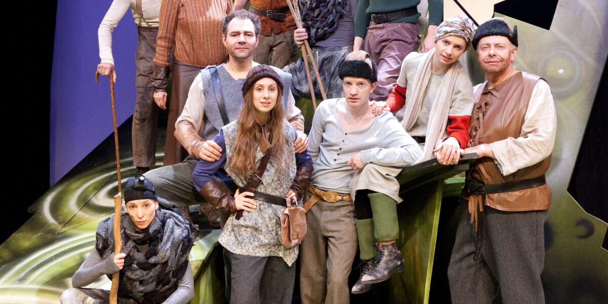 Eine Gruppe von Schauspielern in mittelalterlichen Kostümen posiert vor einer stilisierten Kulisse mit grünen und schwarzen abstrakten Formen, die an eine Theateraufführung erinnern.