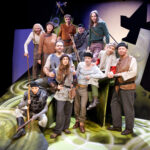 Eine Gruppe von Schauspielern in mittelalterlichen Kostümen posiert vor einer stilisierten Kulisse mit grünen und schwarzen abstrakten Formen, die an eine Theateraufführung erinnern.