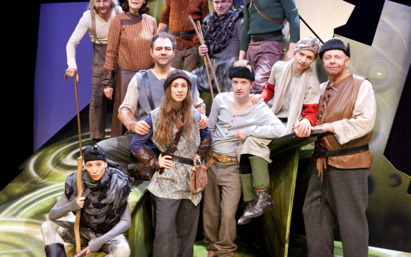 Eine Gruppe von Schauspielern in mittelalterlichen Kostümen posiert vor einer stilisierten Kulisse mit grünen und schwarzen abstrakten Formen, die an eine Theateraufführung erinnern.