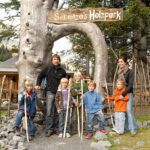 Eine Gruppe von zwei Erwachsenen und fünf Kindern steht unter einem hölzernen Torbogen mit dem Schild "Schnitza's Holzpark", jeder hält einen Spazierstock.