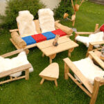Eine Sitzgarnitur aus Holz mit vier Stühlen und einem Tisch auf Gras, mit weißen Pelzüberwürfen und roten oder blauen Sitzkissen.
