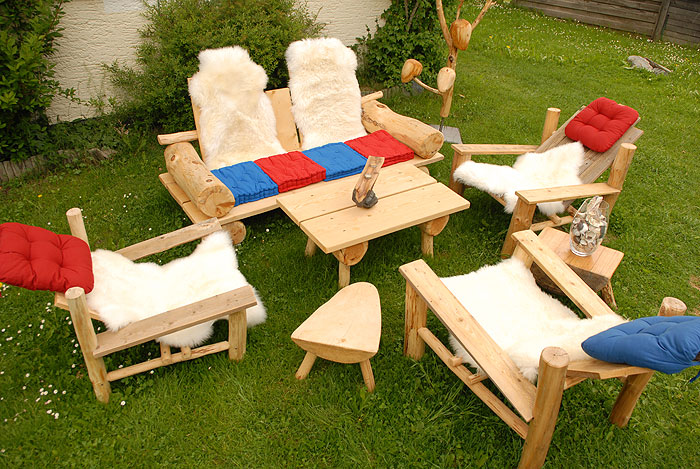 Eine Sitzgarnitur aus Holz mit vier Stühlen und einem Tisch auf Gras, mit weißen Pelzüberwürfen und roten oder blauen Sitzkissen.