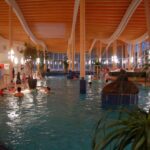 Hallenbad mit Menschen, die schwimmen und sich entspannen; Holzbalken an der Decke, große Fenster, Topfpflanzen und künstliche Beleuchtung sind zu sehen.