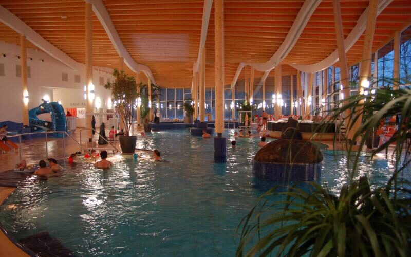 Hallenbad mit Menschen, die schwimmen und sich entspannen; Holzbalken an der Decke, große Fenster, Topfpflanzen und künstliche Beleuchtung sind zu sehen.