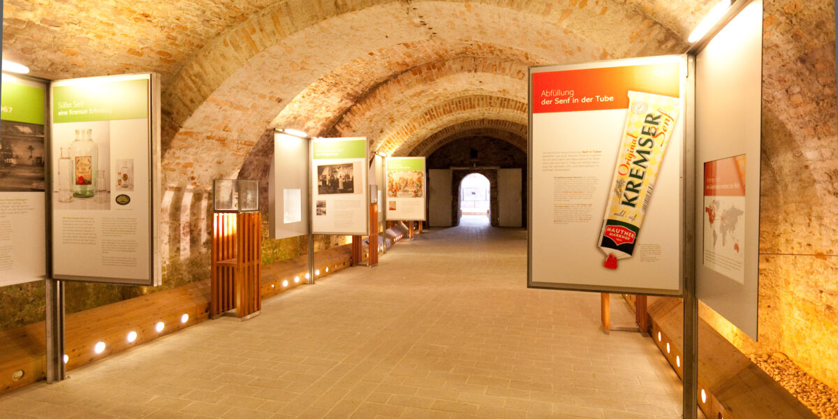 In einem Flur mit Ziegelgewölbe sind Informationstafeln und Bilder zu sehen, darunter das große Bild einer Senftube, in einem Museum oder einer Ausstellung.