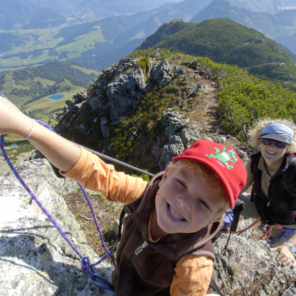 Ein Kind und ein Erwachsener, die Rucksäcke und Klettergurte tragen, klettern an einem Sicherungsseil auf einen felsigen Bergrücken, unter dem ein malerisches Tal zu sehen ist.