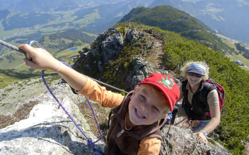 Ein Kind und ein Erwachsener, die Rucksäcke und Klettergurte tragen, klettern an einem Sicherungsseil auf einen felsigen Bergrücken, unter dem ein malerisches Tal zu sehen ist.