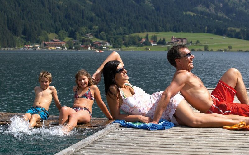 Eine vierköpfige Familie entspannt sich auf einem hölzernen Steg an einem See; zwei Kinder sitzen mit den Füßen im Wasser, während zwei Erwachsene in Badeanzügen vor dem Hintergrund der Berge und Bäume faulenzen.
