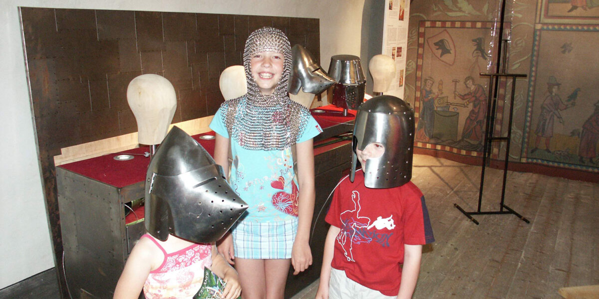 Drei Kinder in einem Museum tragen mittelalterliche Helme, eines davon zusätzlich ein Kettenhemd. Im Hintergrund sind historische Schautafeln und Bildtafeln zu sehen.