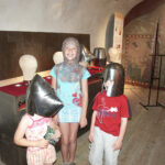 Drei Kinder in einem Museum tragen mittelalterliche Helme, eines davon zusätzlich ein Kettenhemd. Im Hintergrund sind historische Schautafeln und Bildtafeln zu sehen.