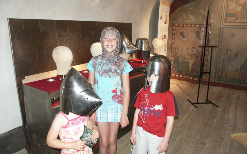 Drei Kinder in einem Museum tragen mittelalterliche Helme, eines davon zusätzlich ein Kettenhemd. Im Hintergrund sind historische Schautafeln und Bildtafeln zu sehen.