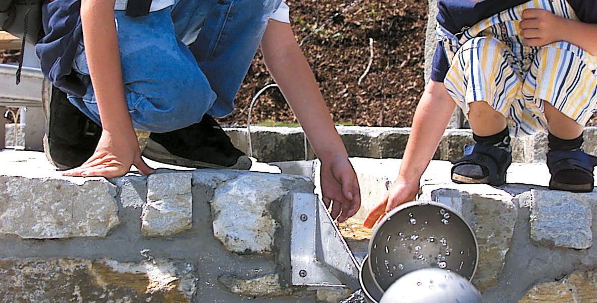 Zwei Kinder knien an einer Steinmauer und fangen mit Metallbechern Wasser auf, das aus einem Springbrunnen im Freien fließt.