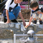 Zwei Kinder knien an einer Steinmauer und fangen mit Metallbechern Wasser auf, das aus einem Springbrunnen im Freien fließt.
