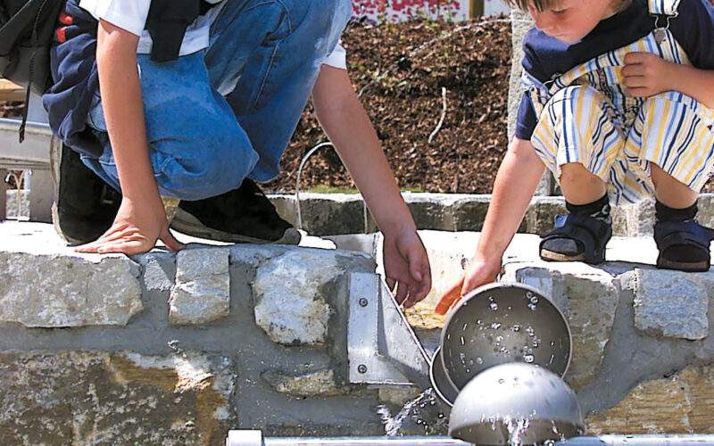 Zwei Kinder knien an einer Steinmauer und fangen mit Metallbechern Wasser auf, das aus einem Springbrunnen im Freien fließt.