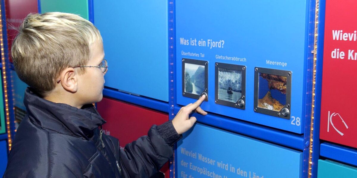 Ein Junge, der eine Brille und eine dunkle Jacke trägt, interagiert mit einem Bildungsdisplay und beantwortet über einen Touchscreen eine Frage über Fjorde auf Deutsch.