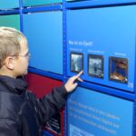 Ein Junge, der eine Brille und eine dunkle Jacke trägt, interagiert mit einem Bildungsdisplay und beantwortet über einen Touchscreen eine Frage über Fjorde auf Deutsch.