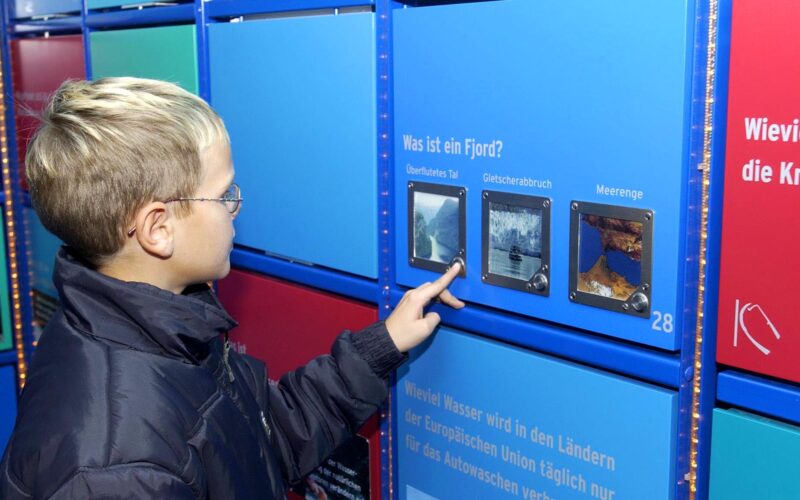 Ein Junge, der eine Brille und eine dunkle Jacke trägt, interagiert mit einem Bildungsdisplay und beantwortet über einen Touchscreen eine Frage über Fjorde auf Deutsch.
