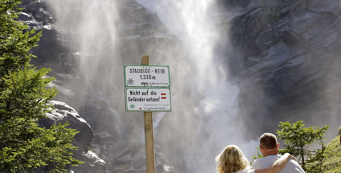 Eine dreiköpfige Familie steht an einem Holzzaun und blickt auf einen großen Wasserfall; ein Schild warnt die Besucher, aus Sicherheitsgründen hinter der Absperrung zu bleiben.