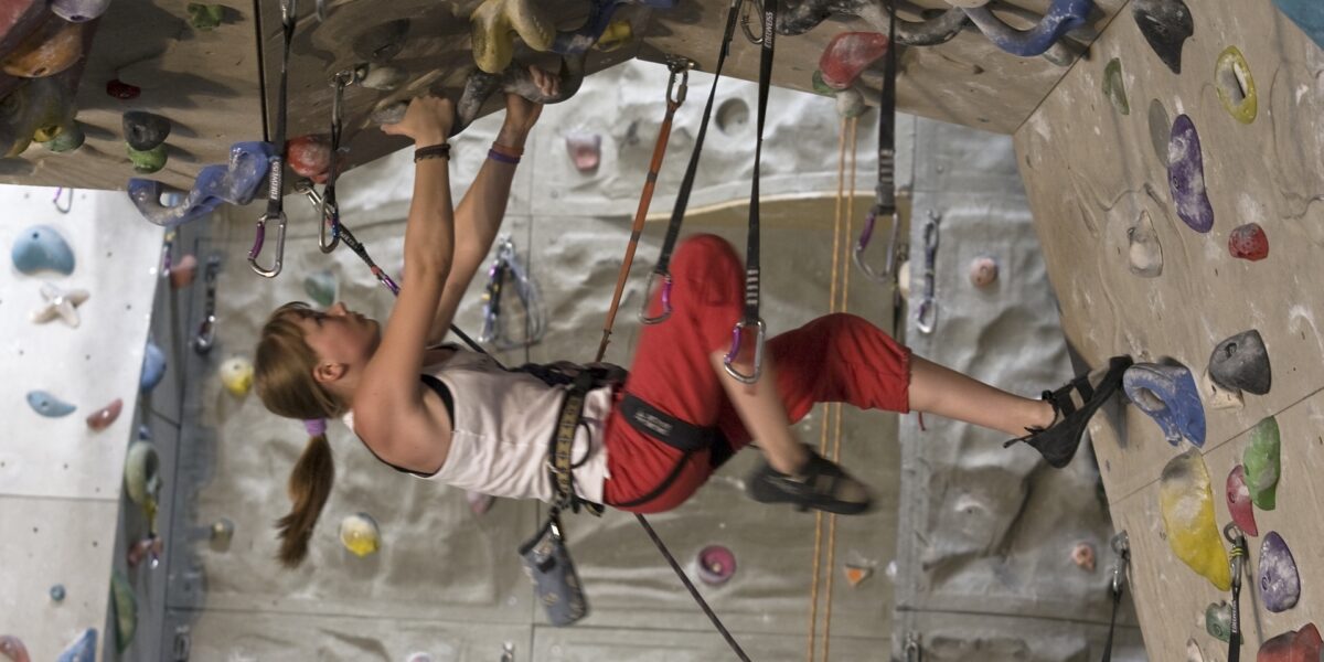 Eine Person mit Klettergurt und Kletterschuhen klettert an einer Indoor-Kletterwand hoch und hält sich an einem überhängenden Abschnitt fest.