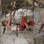 Eine Person mit Klettergurt und Kletterschuhen klettert an einer Indoor-Kletterwand hoch und hält sich an einem überhängenden Abschnitt fest.