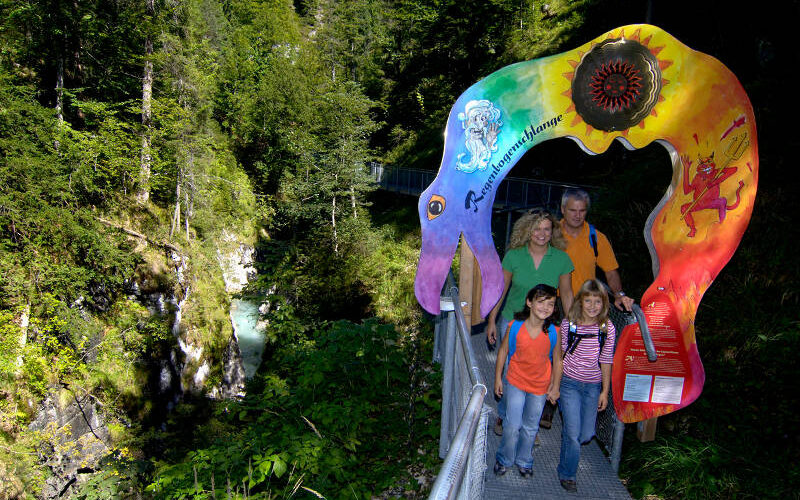 Vier Personen stehen auf einem Metallsteg in einer bewaldeten Schlucht und gehen unter einem farbenfrohen, kunstvollen Torbogen mit Text und Symbolen hindurch.