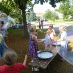 An einem sonnigen Tag spielen Kinder im Freien unter Bäumen und machen große Seifenblasen mit Stäben und Schalen mit Seifenblasenlösung.