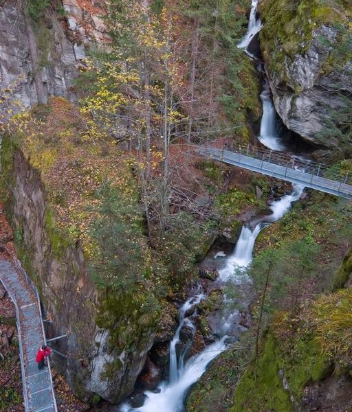 Erhöhte Metallstege überspannen eine enge Schlucht mit Wasserfällen, umgeben von Bäumen und moosbewachsenen Felsen; eine Person in einer roten Jacke geht auf dem Weg.