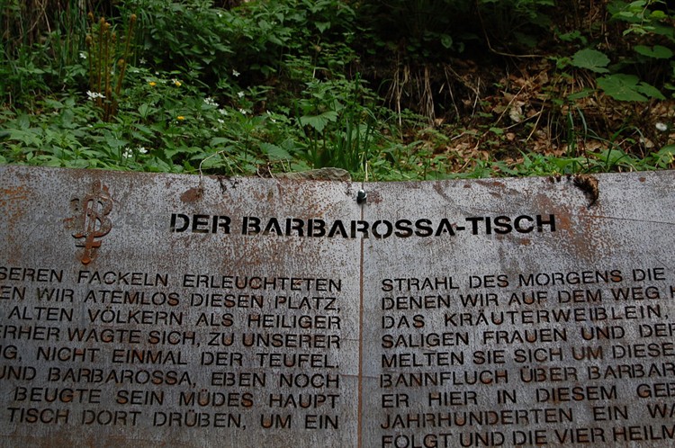 Ein verwittertes Metallschild in deutscher Sprache mit der Aufschrift "DER BARBAROSSA-TISCH", teilweise umgeben von grünen Pflanzen und Blättern.