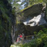 Drei Personen stehen auf einer hölzernen Plattform unter einem großen Felsüberhang in einer üppigen, bewaldeten Gegend mit Moos und Bäumen.