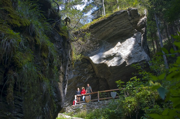 Drei Personen stehen auf einer hölzernen Plattform unter einem großen Felsüberhang in einer üppigen, bewaldeten Gegend mit Moos und Bäumen.