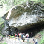 Eine Gruppe von Menschen in traditioneller Kleidung versammelt sich unter einem großen Felsüberhang in einem Waldgebiet, in dessen Nähe Baumstämme und aufgeschichtete Steine liegen.