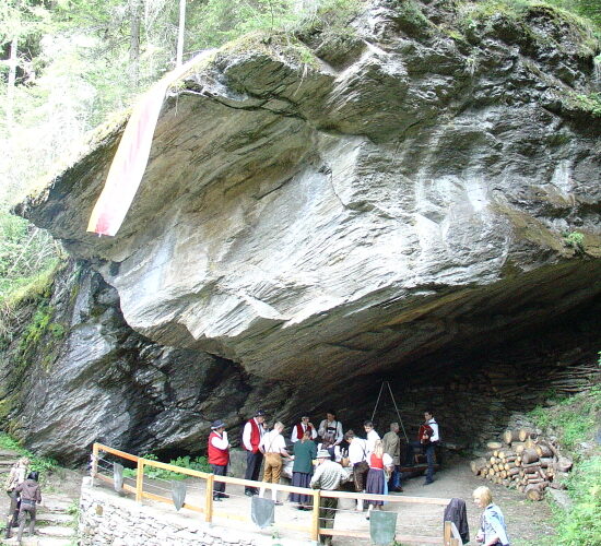 Eine Gruppe von Menschen in traditioneller Kleidung versammelt sich unter einem großen Felsüberhang in einem Waldgebiet, in dessen Nähe Baumstämme und aufgeschichtete Steine liegen.