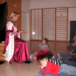 Eine Frau in einem langen, rosafarbenen Kleid sitzt und gestikuliert, während sie zu einer Gruppe von Kindern spricht, die in einem Klassenzimmer auf Matten sitzen.