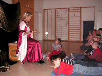 Eine Frau in einem langen, rosafarbenen Kleid sitzt und gestikuliert, während sie zu einer Gruppe von Kindern spricht, die in einem Klassenzimmer auf Matten sitzen.