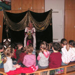 Eine Frau im Kostüm sitzt auf einer Bühne mit drapiertem Hintergrund und liest einer Gruppe kleiner Kinder vor, die auf Bänken ihr gegenüber sitzen.