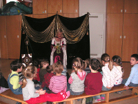 Eine Frau im Kostüm sitzt auf einer Bühne mit drapiertem Hintergrund und liest einer Gruppe kleiner Kinder vor, die auf Bänken ihr gegenüber sitzen.