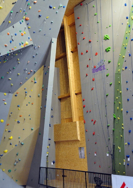 Kletterhalle mit hohen künstlichen Kletterwänden, mehrfarbigen Griffen, Seilen und einem schwarzen Metallgeländer mit einem einzelnen Stuhl im Vordergrund.